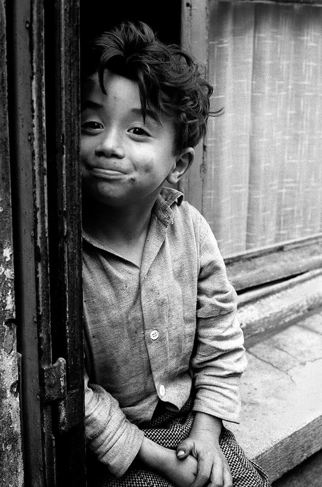 Schwarzweißfoto eines lächelnden Jungen, der im Türrahmen sitzt und die Hände gefaltet hält.